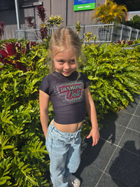 Child wearing Diamond Doll Baby Tee with stylish graphics and fitted design, surrounded by greenery.