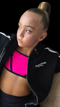 Woman wearing a black jacket with 'Diamond Doll' branding, pink sports bra, and black leggings on a black background.