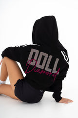 Person wearing a black hoodie with 'DOLL' text, sitting on a white background
